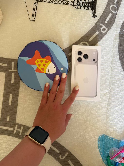 A hand with a white Apple Watch rests on a road-themed play mat next to an iPhone box and a round cushion featuring a cartoon fox design.