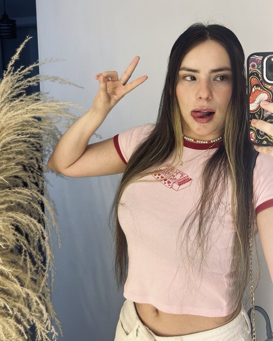 A young woman with long dark hair taking a mirror selfie in a pink ringer t-shirt, making a peace sign with her hand.