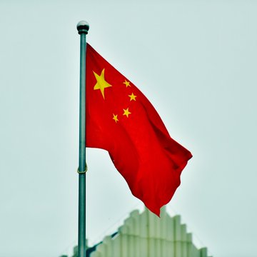 The Chinese national flag, with its distinctive red field and five yellow stars, waves against a hazy sky. The image visually underscores the post's news about Chinese prosecutors pushing for stricter anti-money laundering measures targeting crypto mixers and privacy coins.