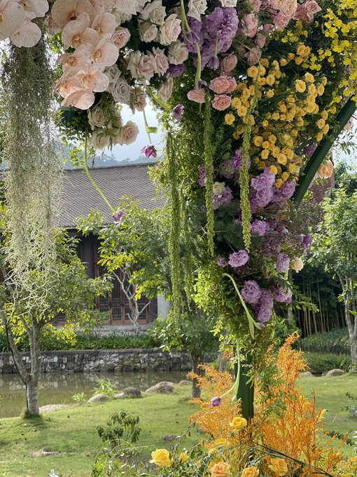 A picturesque garden scene featuring elaborate floral arrangements with pink, yellow, and purple flowers hanging from above, with a traditional building and small pond visible in the background.