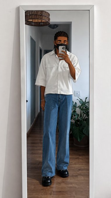Mirror selfie of a person with long braided hair wearing a white button-up shirt and loose blue jeans, standing in a hallway with a plant nearby.