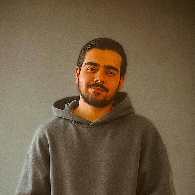 A portrait photograph of a young man with dark hair and a beard, wearing a gray hoodie against a plain gray wall.