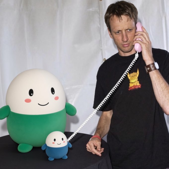 A man wearing a black graphic t-shirt holds a pink telephone receiver to his ear, looking skeptically at two round mascot characters (one large green-and-white, one small blue-and-white) on a table.