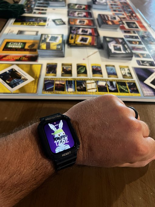 A person's wrist wearing a black "PROTECTION" and "RESIST" smartwatch displaying a character and the time 12:52, with a Star Wars Unlimited card game in progress on the table behind it.