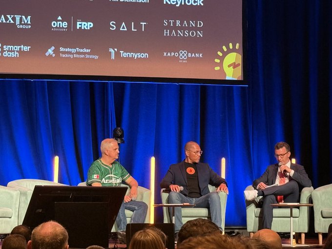 A man wearing a green shirt with an orange logo sitting between two other men at a conference.