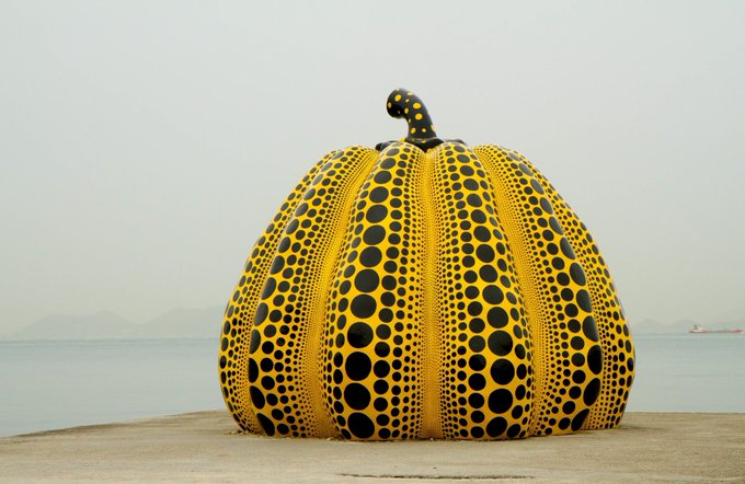 A large yellow pumpkin sculpture covered in black polka dots, displayed outdoors by the sea, characteristic of artist Yayoi Kusama.
