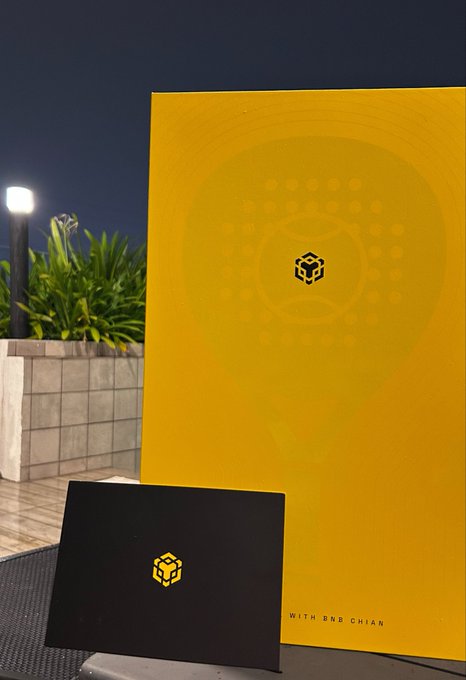 A bright yellow folder with a subtle face design and the text 'WITH BNB CHIAN' sits next to a black card with a matching cube logo on an outdoor table at night.