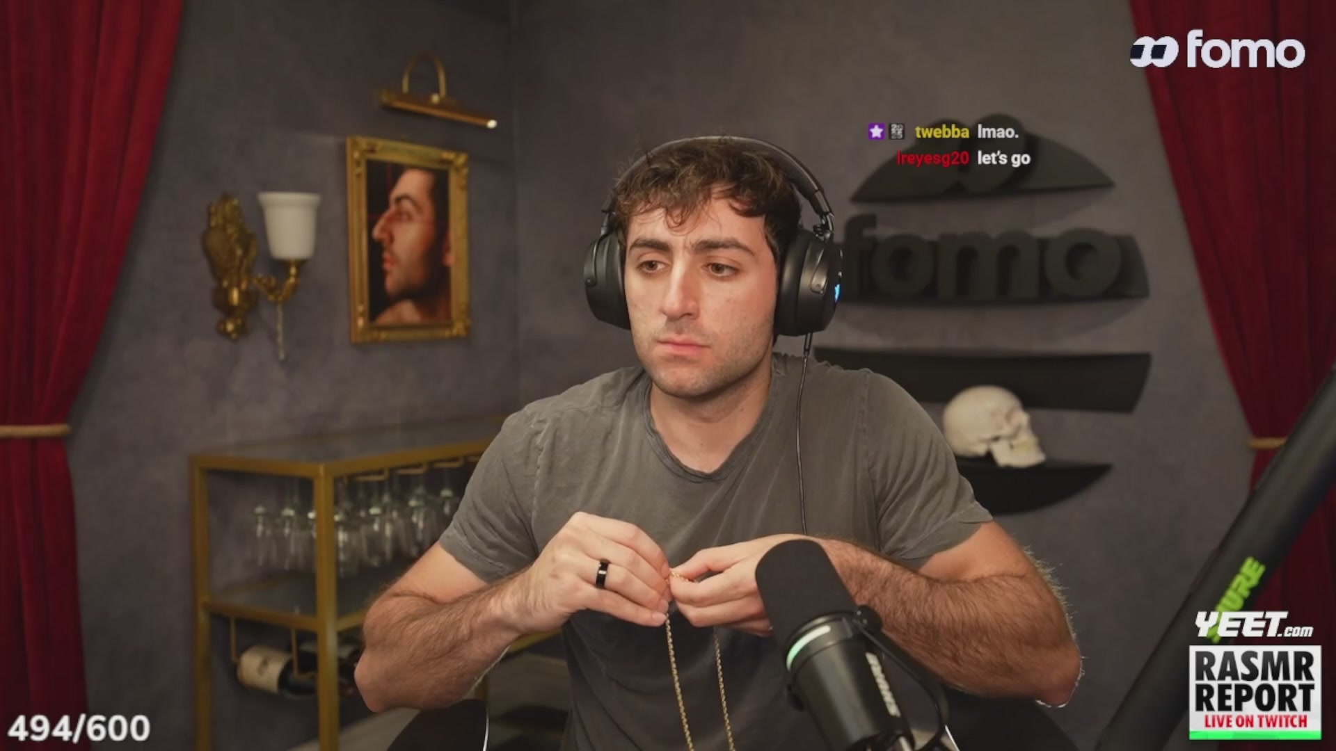 urn of gold and white flowers, framed picture on wall, black curtain with gold trim, man in gray t-shirt and headphones, green microphone stand, green and white logo, "FOMO" text, and a red curtain backdrop.