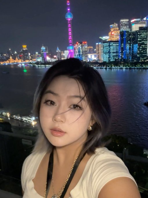 A selfie of a young woman with short hair in front of the illuminated Shanghai skyline at night, with the Oriental Pearl Tower visible behind her.