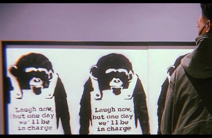 A Banksy-style artwork or photograph showing the iconic 'Laugh now, but one day we'll be in charge' monkey stencil print displayed on a wall, with a person in a green jacket partially visible on the right side.