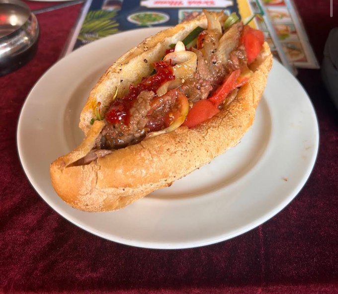 A Vietnamese bánh mì sandwich filled with grilled meat, pickled vegetables, and red chili sauce sits on a white plate atop a maroon tablecloth. The accompanying post about frustration with crypto wallet efforts ("retro coin zksync") seems unrelated to the image, suggesting the food may simply be a meal shared during a moment of reflection or venting.