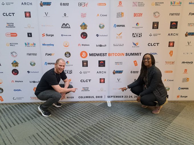 Two people crouching and pointing at each other in front of a step-and-repeat backdrop for the 'MIDWEST BITCOIN SUMMIT' held in Columbus, Ohio on September 23-24, 2025, featuring sponsor logos like ARCH, BitPay, Bitcoin Well, and CLCT.