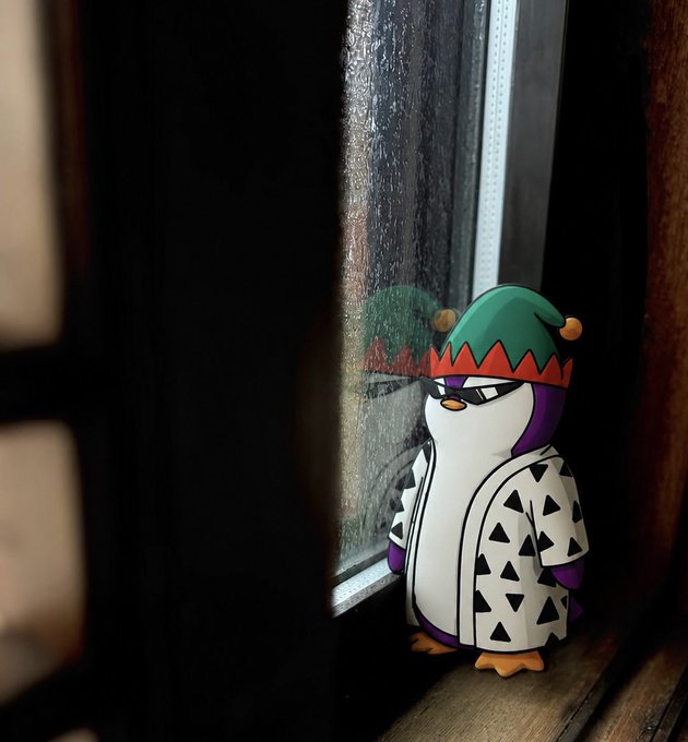 A cartoon penguin figure wearing a green elf hat, sunglasses, and a white shirt with black triangles, standing on a wooden windowsill.