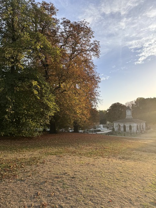 An autumnal park scene featuring a large tree with orange and green leaves beside a pond, with a small classical-style pavilion in the background under a partly cloudy sky.