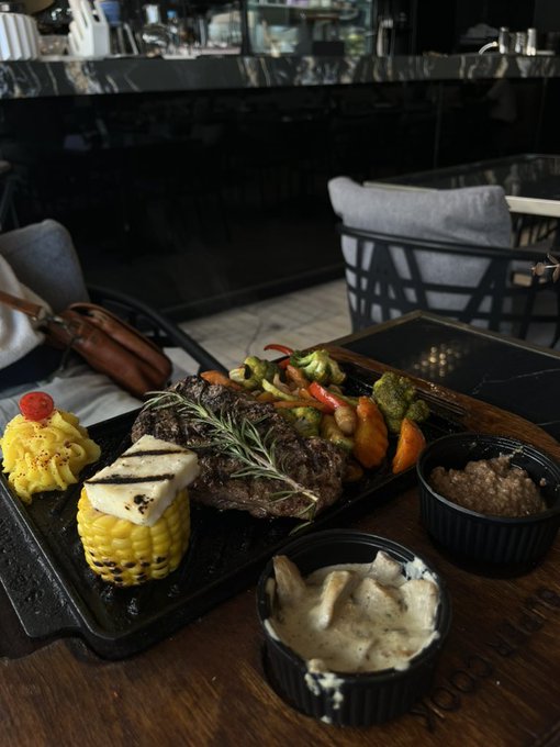 A restaurant meal is served on a black cast iron platter, featuring a grilled steak garnished with rosemary, corn on the cob, grilled vegetables, and a cherry tomato, accompanied by two small ramekins of side dishes on a wooden table.