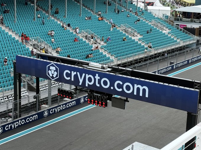The image shows an empty race track with a blue sign that reads "@cryptocom" above it, indicating that this is a Cryptomining event. The sign also mentions that there are exclusive passes available for the @automotive GM As the Formula 1 season heats up with high-speed battles and roaring engines. The text suggests that attendees can win a prize by participating in the event.