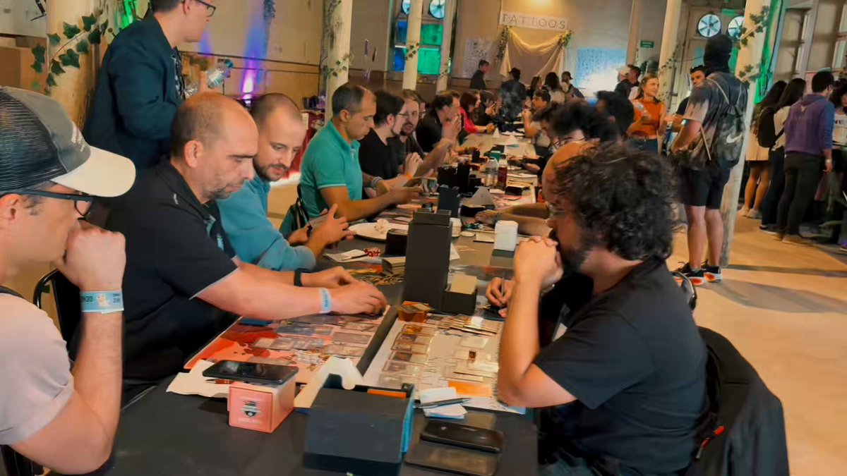 The image shows a group of people sitting around tables, playing a video game together. There are at least 12 people visible in the scene, with some players actively engaged in the game while others watch or wait for their turn. A few individuals can be seen wearing hats and backpacks, indicating that they might have come from different places to participate in this gaming event. The tables are covered with various items such as cards, dice, and other game components, suggesting a competitive atmosphere among the players.