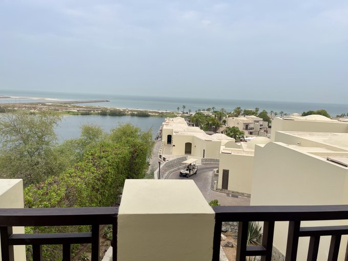 A balcony view overlooking a coastal resort with white Mediterranean-style buildings, palm trees, a lagoon, and the ocean in the distance.