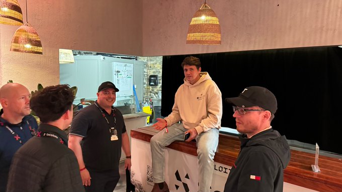 A casually dressed man in a cream Nike hoodie sits on a counter in a cafe, gesturing while talking to three other men wearing polo shirts and a Tommy Hilfiger jacket.