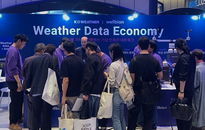 A busy trade show booth for 'KO WEATHER | wellbian' promoting 'Weather Data Economy' in Korean, with numerous attendees in business attire gathered around displays of weather monitoring equipment.