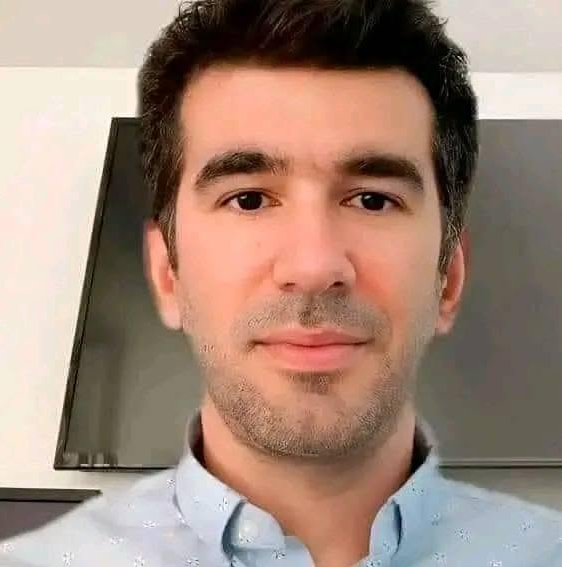 A selfie-style portrait of a man with dark hair and stubble wearing a light blue patterned button-up shirt, looking directly at the camera with a subtle smile. The humorous caption suggesting modest financial hopes of "a few hundred million" plays on the contrast between his casual, approachable appearance and the exaggerated ambition of the statement.