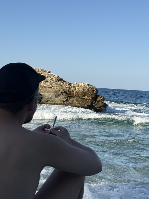 Over-the-shoulder shot of a shirtless man in a dark cap and sunglasses using his phone on a beach, with rocky cliffs and ocean waves in the background.