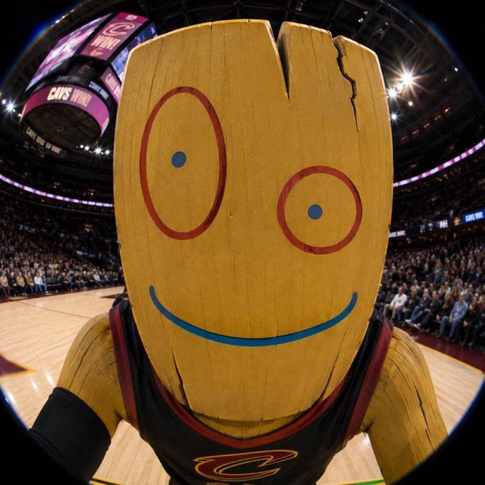 The Cleveland Cavaliers mascot, a wooden plank with a smiley face and red circles around the eyes, photographed with a fisheye lens in an arena with 'CAVS WIN!' visible on the scoreboard.