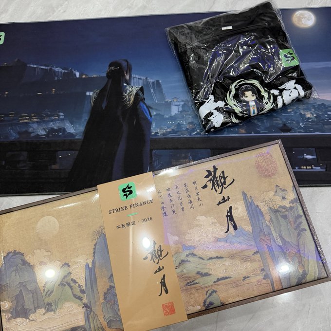 A photograph of a 'Strike Finance 2026' gift box featuring traditional Chinese landscape artwork with the text '觀山月' (Guan Shan Yue), alongside a black plastic-wrapped item with anime character designs.