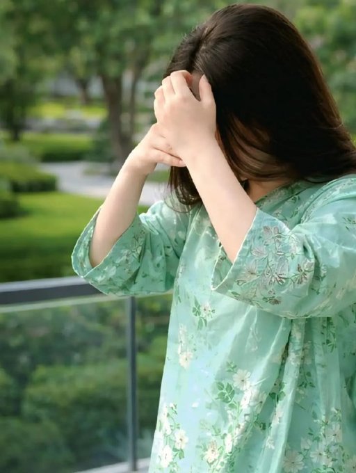 A woman in a flowing mint-green floral dress stands on a balcony, her hands raised to cover her face as she looks out over a green park.