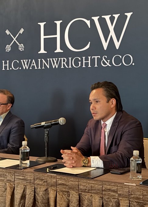 A man in a dark suit and pink tie speaks at a panel discussion against a backdrop displaying "HCW" and "H.C. Wainwright & Co." He is seated at a table with a microphone, water bottles, and notes, appearing to address institutional investors about Bitcoin as collateral, consistent with the post's mention of speaking at an H.C. Wainwright event regarding Strive ($ASST, $SATA).