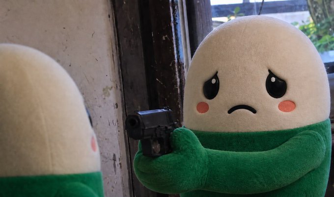 A plush toy with a sad face and green body holds a black handgun, seen reflected in a mirror.