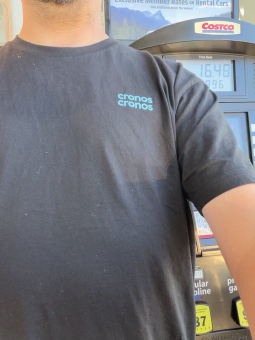 A person wearing a black "cronos" branded t-shirt stands at a Costco gas pump displaying a sale total of $16.96, visually reinforcing the joke about Cronos Network's blockchain transaction fees being lower than Costco's real-world gas prices.