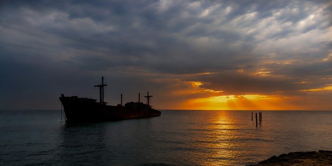 A silhouette of a shipwreck sits in the ocean during a dramatic sunset with sunrays piercing through the clouds.