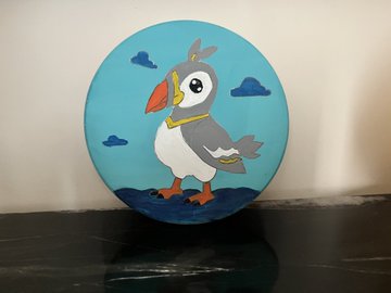 A circular painted artwork depicting a cartoon puffin wearing a gold chain, standing against a blue sky with clouds and water.