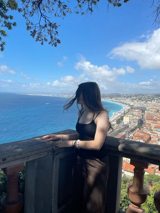 The image features a woman standing near a railing, overlooking a beautiful view of the ocean from an elevated position. She appears to be enjoying her time at this location, possibly taking a break or contemplating something. The sky above her is blue with some clouds scattered across it, adding to the picturesque scenery. In the background, there are buildings and trees that contribute to the overall atmosphere of the scene. The image also contains text elements such as "early morning web3 good Morning @base Family good Morning @Polymarket Builder Creator Active Flw Me".