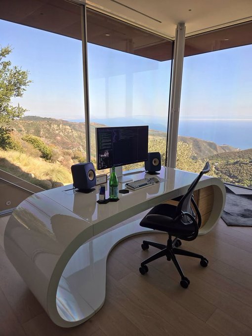 A modern home office setup with a sleek white curved desk, computer monitor, speakers, and an ergonomic chair, overlooking a scenic mountain and ocean view through floor-to-ceiling windows.