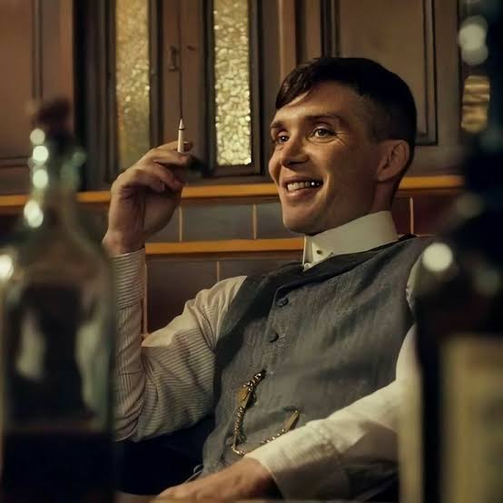 The image shows Cillian Murphy as Tommy Shelby from "Peaky Blinders," sitting in a dimly lit pub, smiling and holding up a cigarette. The caption pairs perfectly with the post's joke about fake optimism, as Shelby's smug, knowing grin mirrors the smugness of investors suddenly claiming they always expected the market recovery.