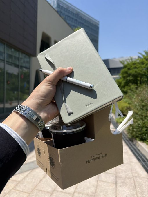 The image shows a person holding a small notebook, pen, and coffee cup outside of an office building. There are two other people visible in the background who appear to be walking by or standing near the building. The scene suggests that someone might have just arrived at work or is taking a break from their daily routine.