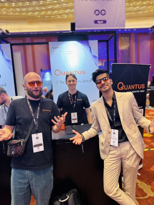 The image shows three men standing next to each other, posing for a photo. They are all wearing name tags that say "Quantum" or "@QuantusNetworks", indicating they might be attending an event related to Quantum computing or cryptocurrency. One of them is holding a bag in his hand. The background features a banner with the words "Quantum" and "@QuantusNetworks".