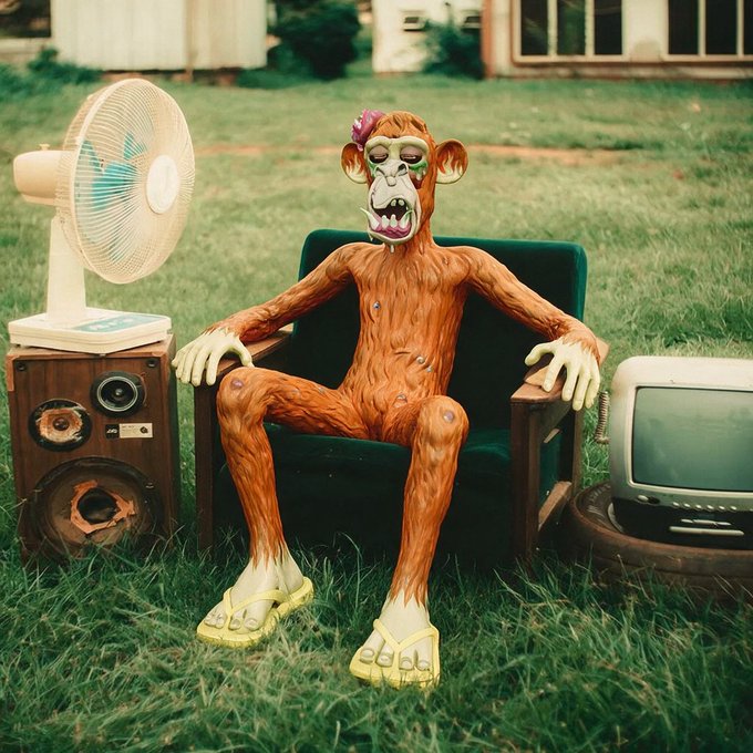 A creepy orange monkey figurine wearing yellow flip-flops and a small bow sits in a green armchair on grass, surrounded by a fan, an old speaker, and a vintage television.