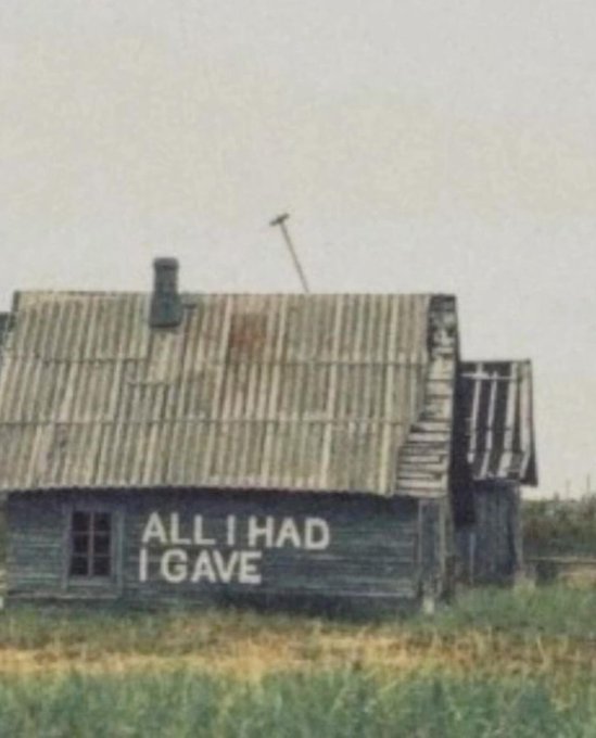 iced glass window on a weathered wooden house, with the words "All I Had I Gave" written in white chalk against a gray sky and grassy field.