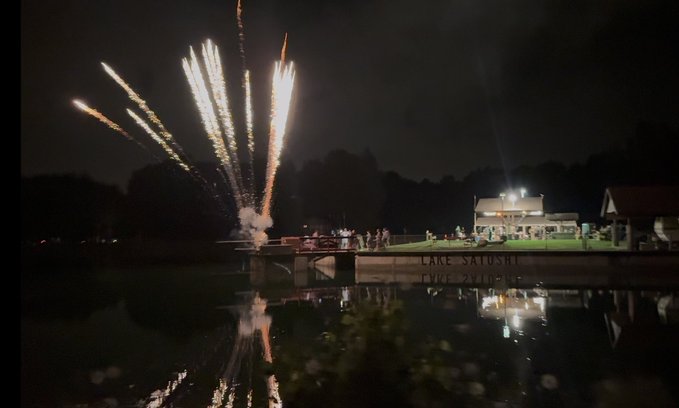 Fireworks explode over Lake Satoshi, casting their colorful reflections across the water as a group of people gather on a lit dock next to a brightly illuminated building. The scene captures the festive first night of an event, matching the caption's description of an enjoyable opening celebration.