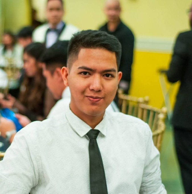 Portrait of a young man with short dark hair, wearing a white patterned shirt and black tie, seated at what appears to be a formal event with other guests behind him.