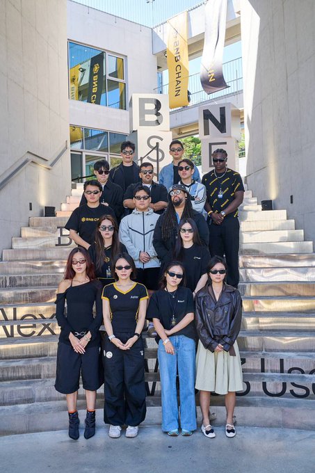 A group of around a dozen people, mostly wearing black or dark clothing, pose on concrete steps outdoors, with large 'BNB' letter sculptures and 'BINANCE' banners visible in the background.