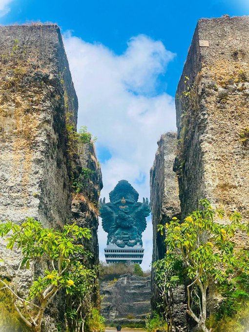 A towering green Garuda statue framed between two massive rock cliffs at GWK Cultural Park in Bali, Indonesia, viewed from below through green foliage against a blue sky with scattered clouds.