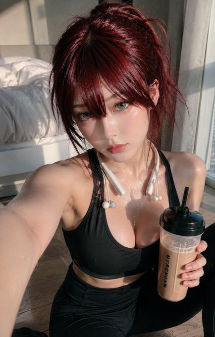 The image features a woman with red hair, wearing black workout clothes while holding a drink. She appears to be taking a selfie or recording a video of herself. The background shows a bedroom setting with a bed visible behind her.