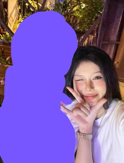 A selfie of a young woman winking and making a peace sign with her hand, wearing a white top and a wristwatch, with a large purple censored/blurred shape covering the left side of the image.