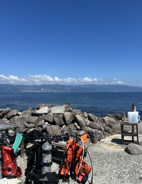 Bicycle touring gear including panniers, bottles, and an orange bike parked on a concrete waterfront overlooking a calm sea with mountains in the distance under a blue sky.