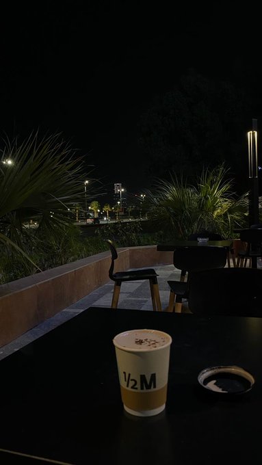 A nighttime outdoor table scene featuring a coffee cup labeled "1/2 M" next to an ashtray, with a patio, chairs, and city lights visible in the background.