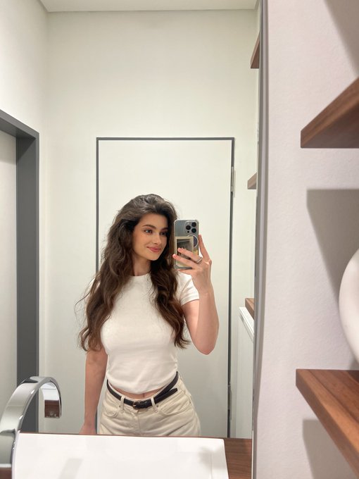 A woman with long brown hair takes a mirror selfie in a bathroom, wearing a white t-shirt and light pants, holding a smartphone.